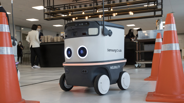An autonomous robot labeled “Samsung C-Lab” and “NEUBILITY” moves indoors between orange safety cones. People are gathered in the background, suggesting a startup showcase or demo event.